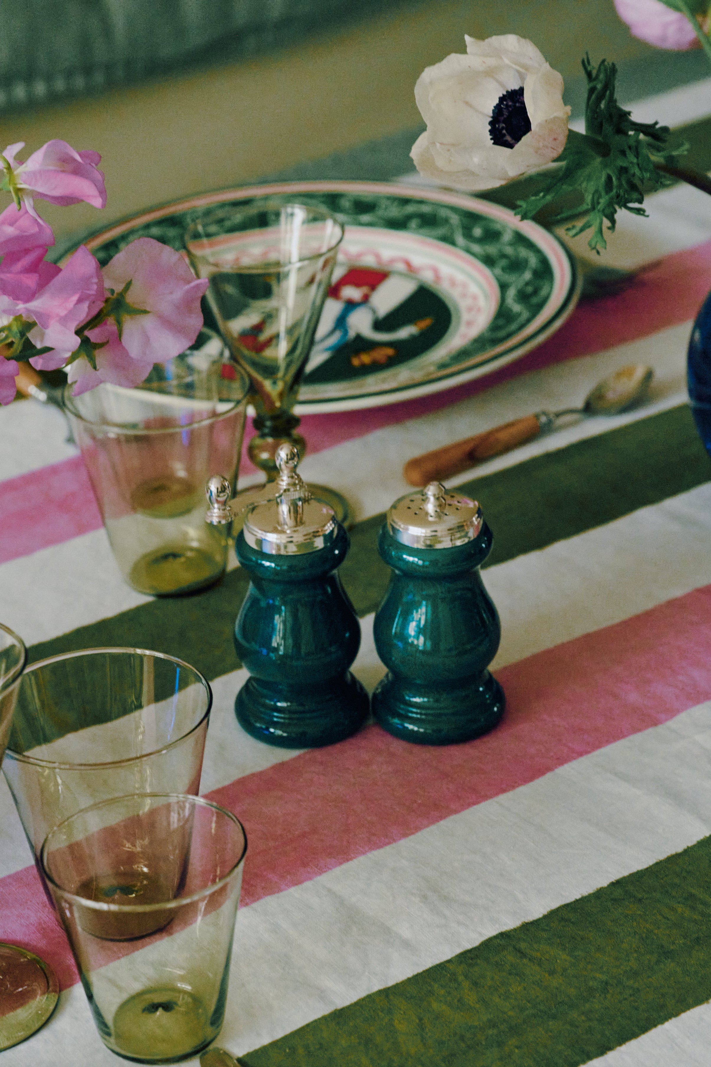 A table set with a striped green, pink, and white tablecloth features glassware, a floral plate, silverware, and the Giorgia Pepper Mill and Salt Shaker—elegantly designed in Italian style with a superior grinding mechanism—beautifully adorned with pink and white flowers in a vase.