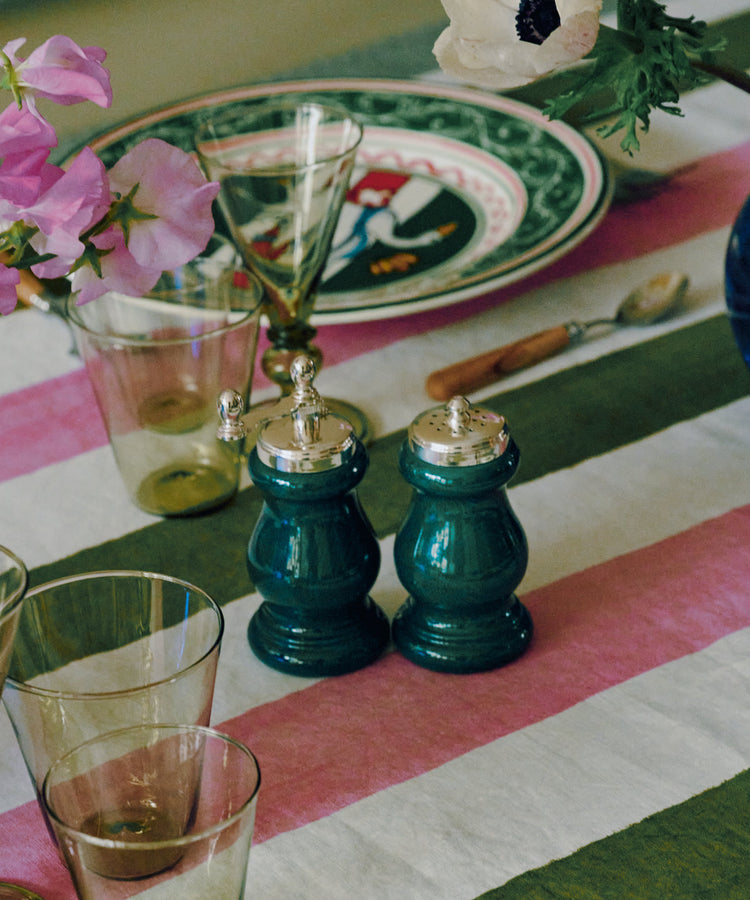 A table set with a striped green, pink, and white tablecloth features glassware, a floral plate, silverware, and the Giorgia Pepper Mill and Salt Shaker—elegantly designed in Italian style with a superior grinding mechanism—beautifully adorned with pink and white flowers in a vase.