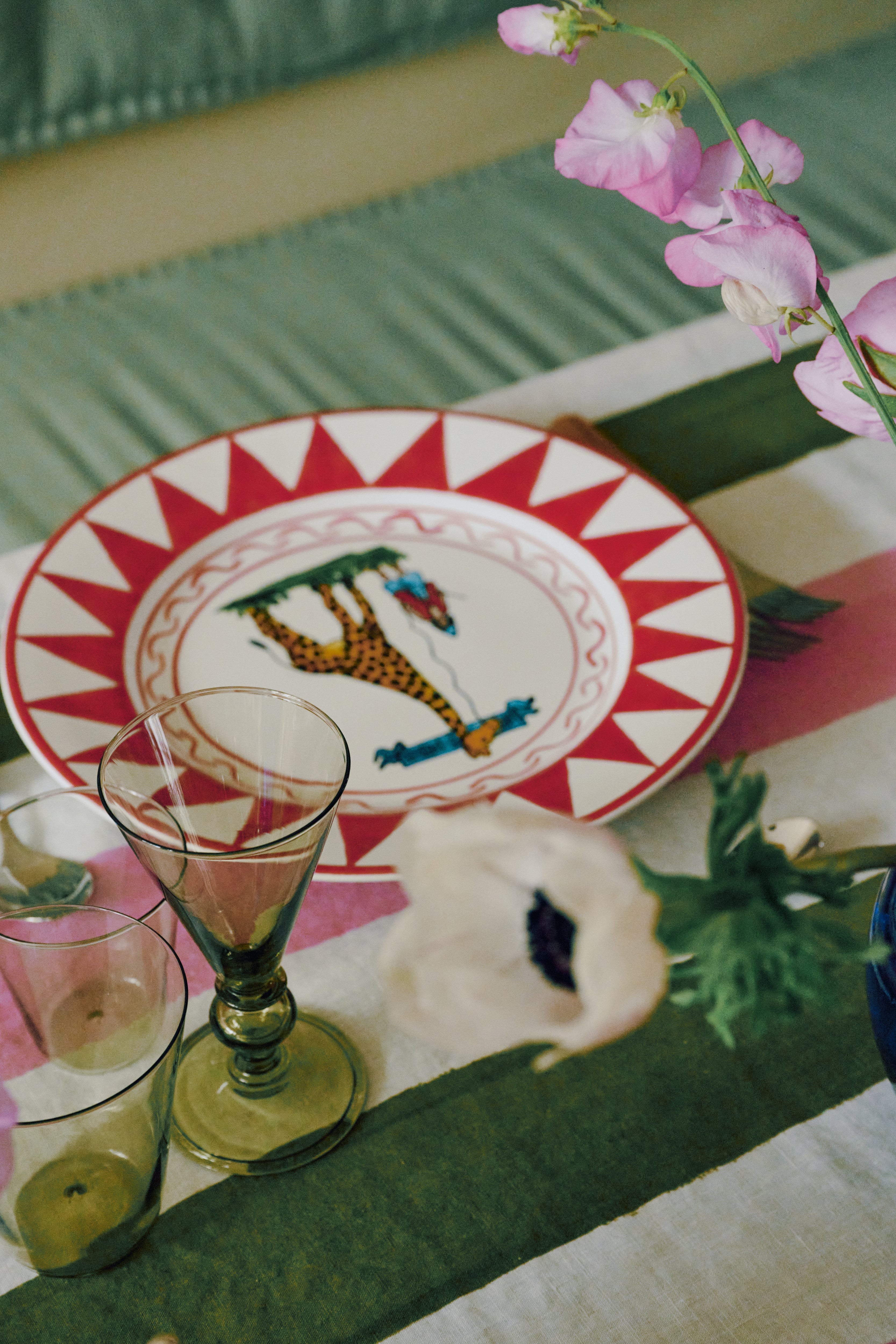 A dinner table set with a Palio Dinner Plate, The Giraffe, two empty glasses, and a striped tablecloth with flowers nearby.