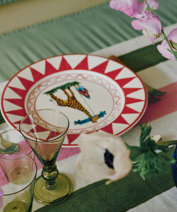A dinner table set with a Palio Dinner Plate, The Giraffe, two empty glasses, and a striped tablecloth with flowers nearby.
