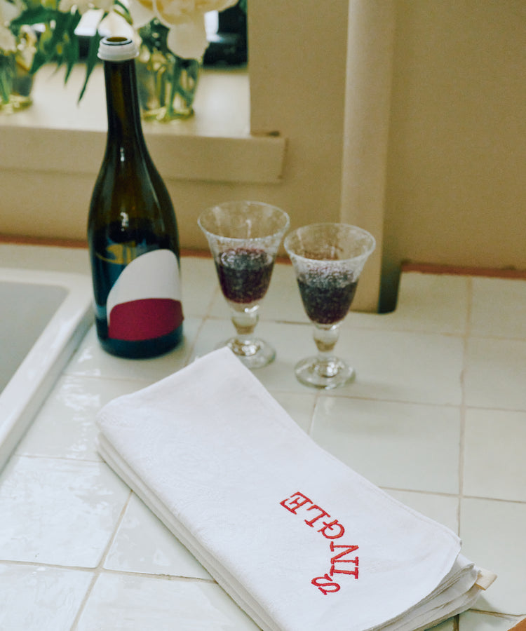 A bottle of wine and two filled glasses sit on a tiled countertop beside the Relationship Status Napkin, Set of 6—one folded with ENTRE in red. Flowers on the windowsill evoke a fun dinner party mood.