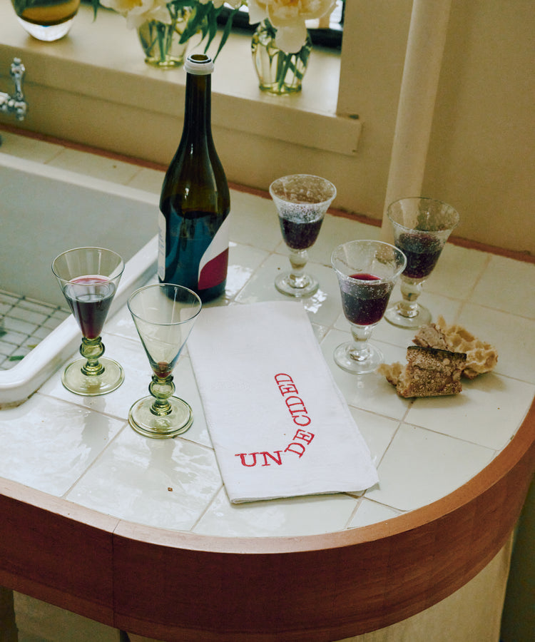 A bottle of red wine, four filled glasses, pieces of bread, and a Relationship Status Napkin from the set of 6 rest on a tiled countertop by a window with flowers.