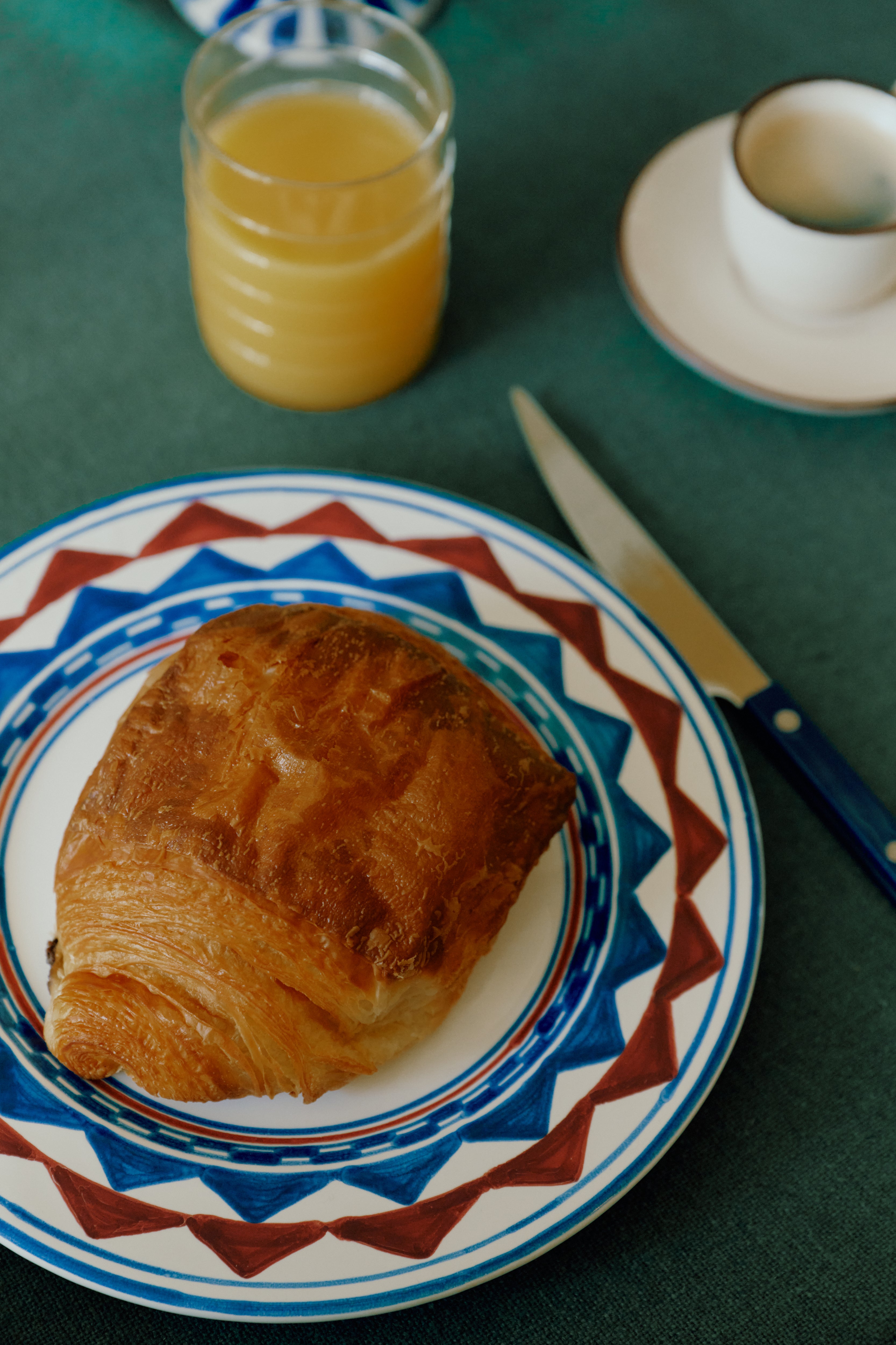 A croissant on a Luca Small Plate, a knife, a glass of orange juice, and a cup of coffee sit atop a green tablecloth.