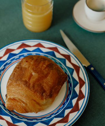 A croissant on a Luca Small Plate, a knife, a glass of orange juice, and a cup of coffee sit atop a green tablecloth.