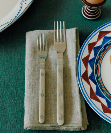 The Bistrot 5 Piece Cutlery Set features two forks resting on a folded napkin, with patterned plates and an egg cup on a green tablecloth for a Parisian bistro-inspired look.