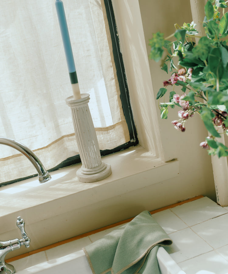 A blue candle in a white holder rests on the kitchen windowsill, with green dish towels, eco-friendly linen placemats and a set of 6 Corte Napkins, plus a nearby vase of flowers.