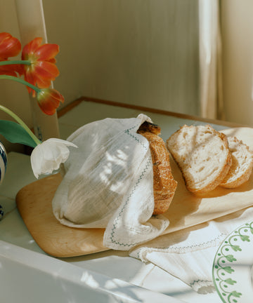A loaf of bread, partially covered with a cloth and three slices cut, rests on the Birds Eye Maple Serving Board beside a vase of red and white flowers.