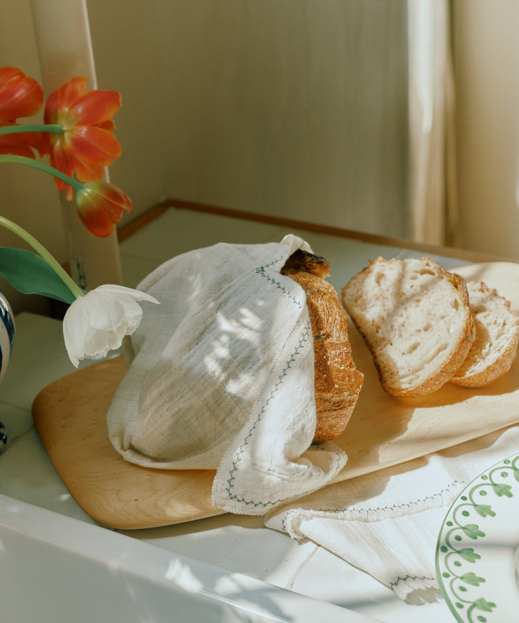 A loaf of bread, partially covered with a cloth and three slices cut, rests on the Birds Eye Maple Serving Board beside a vase of red and white flowers.