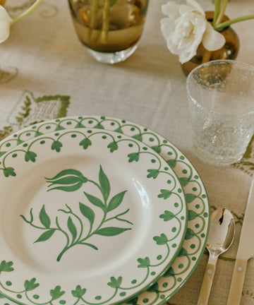 A table setting with two stacked Esmeralda Dinner Plates featuring green floral designs, a clear textured glass, knife, and fork on a patterned tablecloth with flowers in vases.