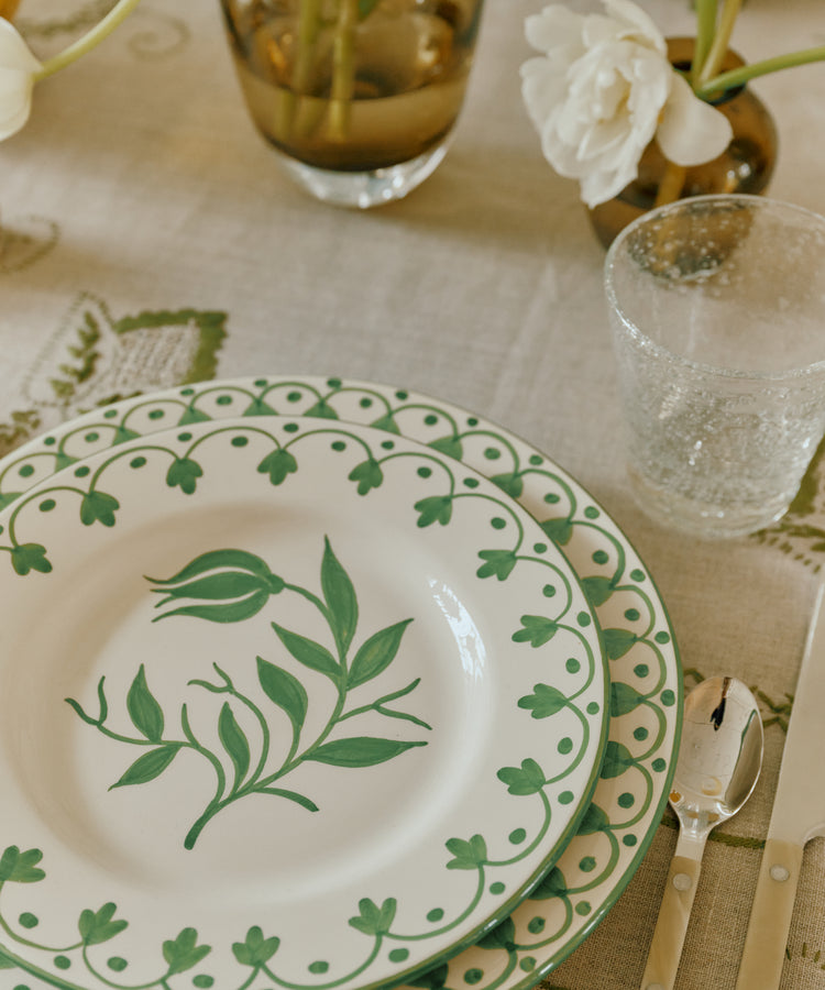 A table setting with two stacked Esmeralda Dinner Plates featuring green floral designs, a clear textured glass, knife, and fork on a patterned tablecloth with flowers in vases.