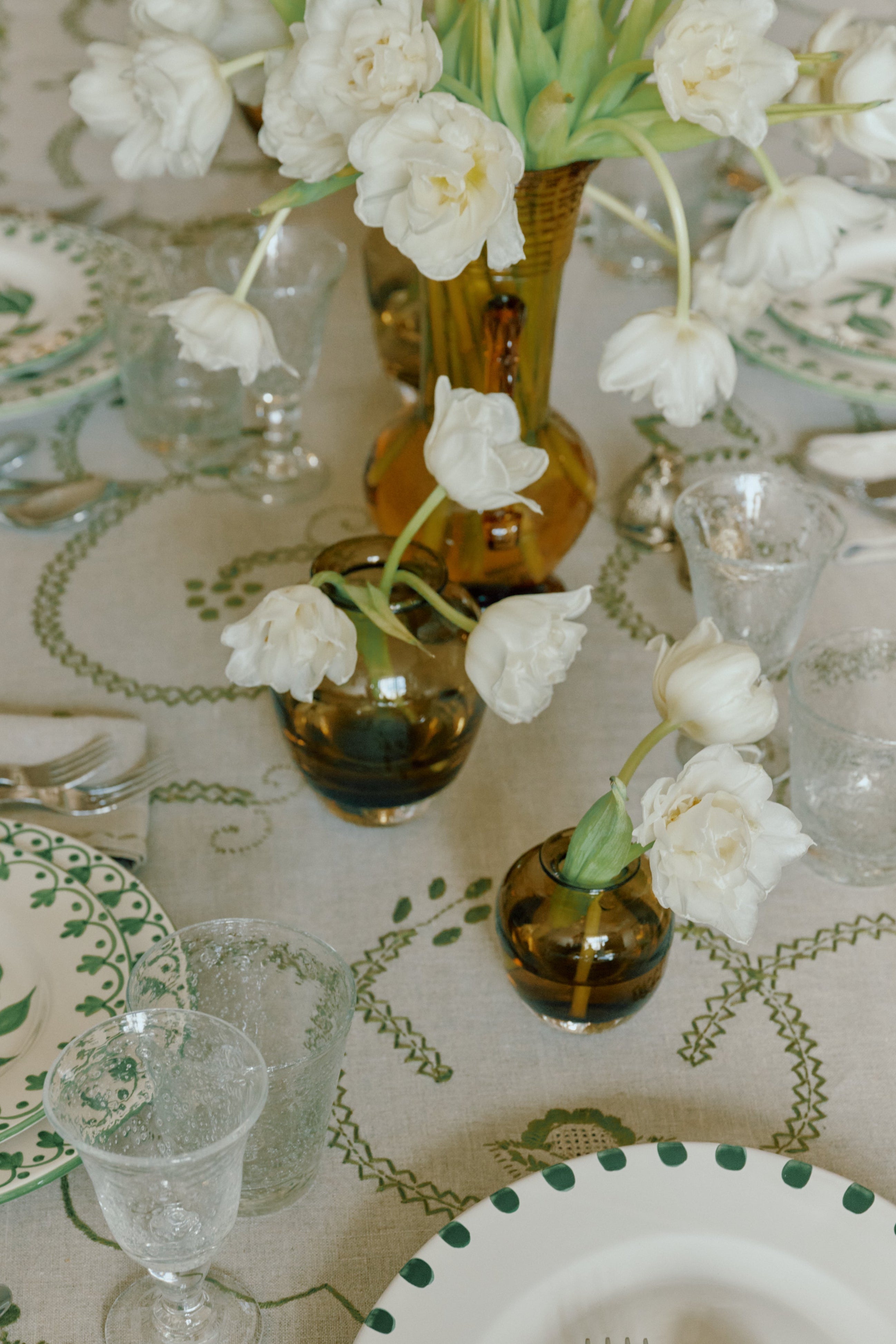 A styled dining table setting with a beige tablecloth featuring a green vine and leaf pattern, white plates with green accents, clear glassware, and white tulips in amber and clear glass vases.