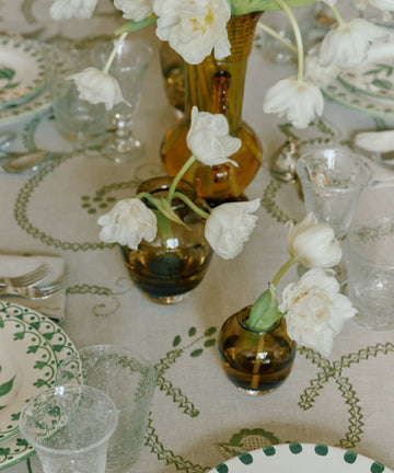 A styled dining table setting with a beige tablecloth featuring a green vine and leaf pattern, white plates with green accents, clear glassware, and white tulips in amber and clear glass vases.