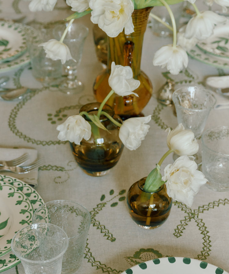 A styled dining table setting with a beige tablecloth featuring a green vine and leaf pattern, white plates with green accents, clear glassware, and white tulips in amber and clear glass vases.