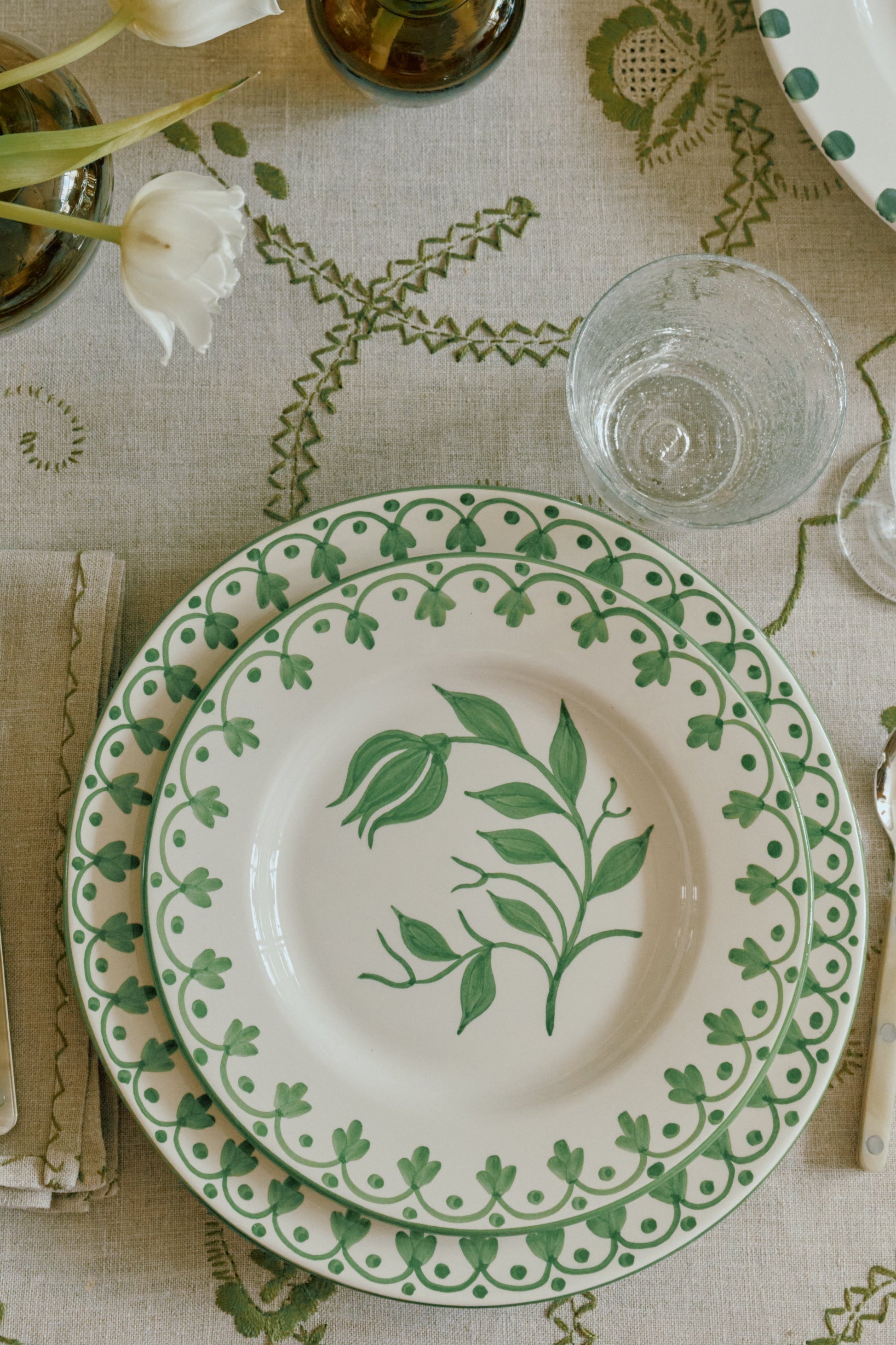 A styled tabletop setting featuring white ceramic plates with green botanical patterns, silver cutlery, clear glass tumblers, and white tulips in small dark glass vases on a beige patterned tablecloth.
