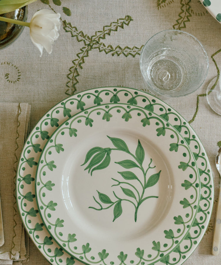 A styled tabletop setting featuring white ceramic plates with green botanical patterns, silver cutlery, clear glass tumblers, and white tulips in small dark glass vases on a beige patterned tablecloth.
