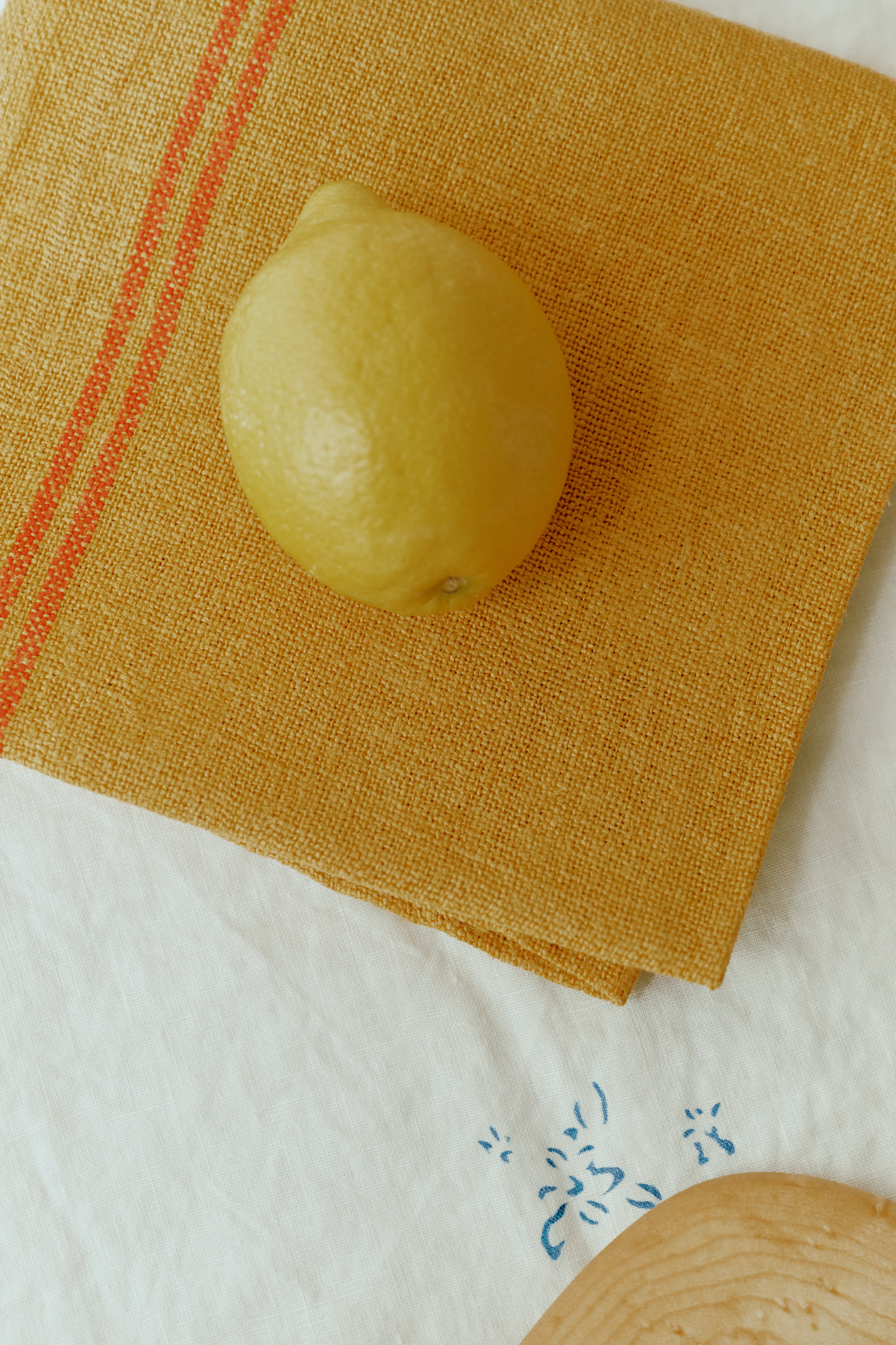 A lemon rests on a French Vintage Tea Towel, richly dyed yellow with an orange stripe, displayed on a white surface with a small blue pattern that evokes the charm of Provence.