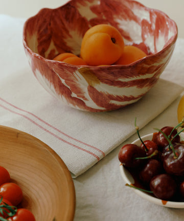 A hand-painted Radicchio Serving Bowl with apricots, a wooden bowl with vine tomatoes, and a small plate of cherries arranged on a white tablecloth.