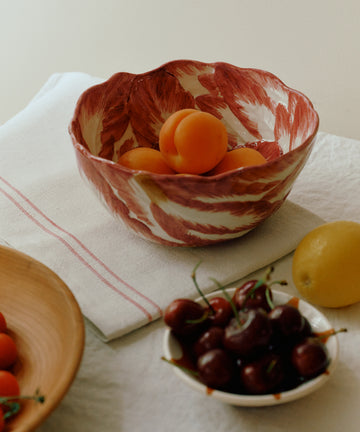 The Radicchio Serving Bowl, hand-painted in red and white, is filled with apricots and rests on a white tablecloth beside a small bowl of cherries and a lemon.