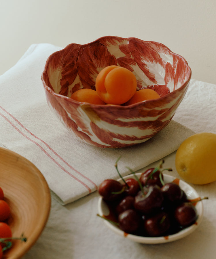 The Radicchio Serving Bowl, hand-painted in red and white, is filled with apricots and rests on a white tablecloth beside a small bowl of cherries and a lemon.