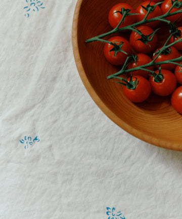A wooden bowl with vine tomatoes rests on the Falling Flower Linen Tablecloth from Summerill & Bishop, adorned with small blue hand-drawn flowers.