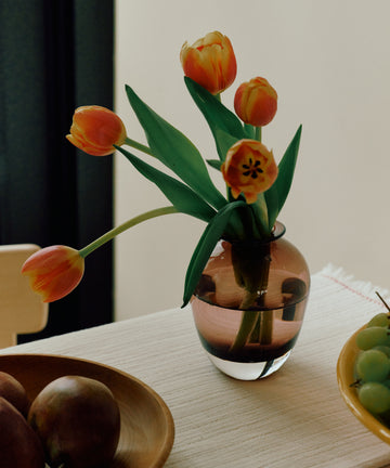 The Gem Bud Vase, hand blown and small, holds orange tulips on a table beside a wooden fruit bowl and a plate of green grapes.