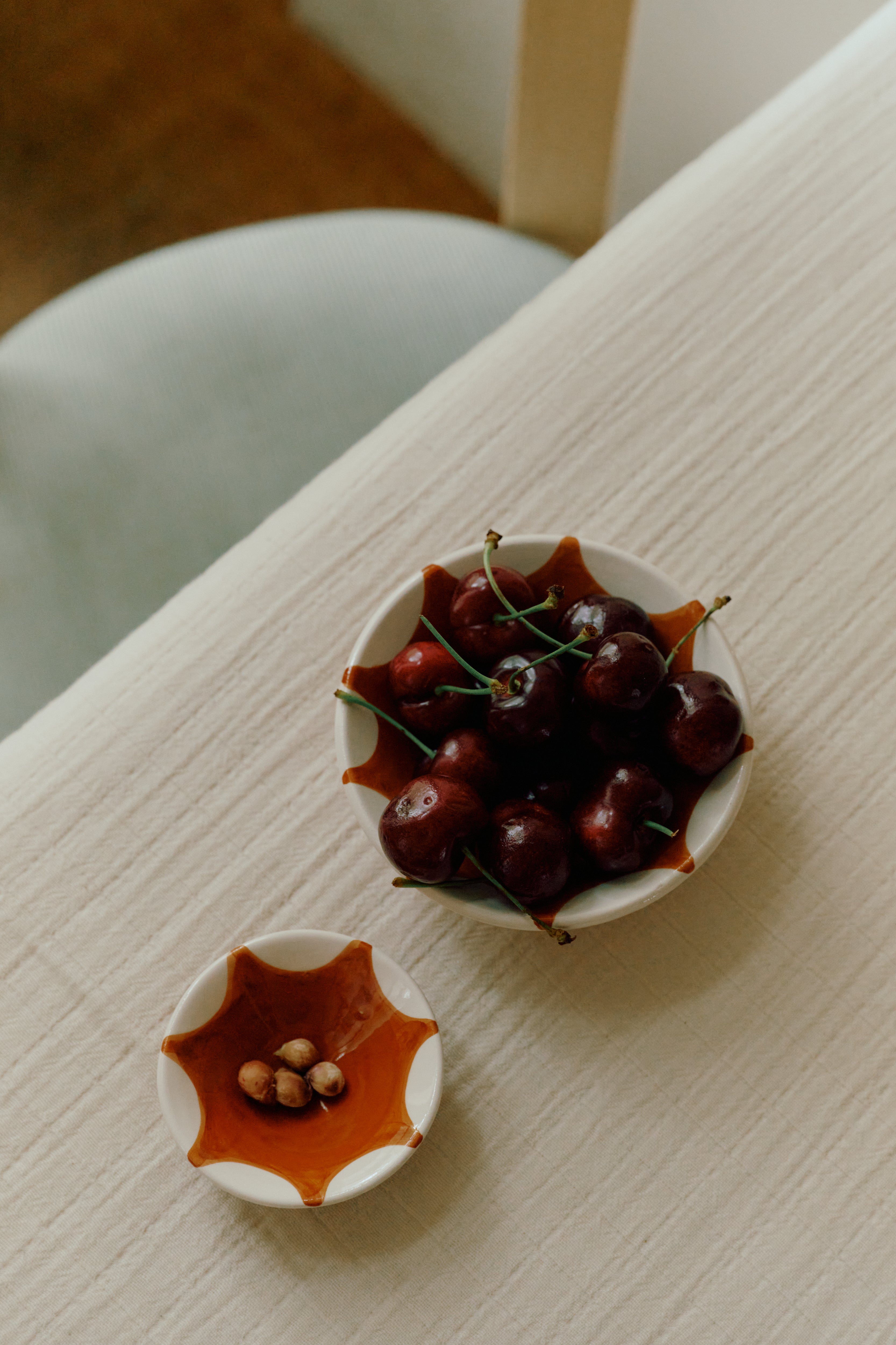 The Stella Nesting Bowls—one filled with cherries and the smaller holding cherry pits—rest on a cream-colored textured tablecloth by a chair, their distinctive ceramics lending artisanal charm to the setting.
