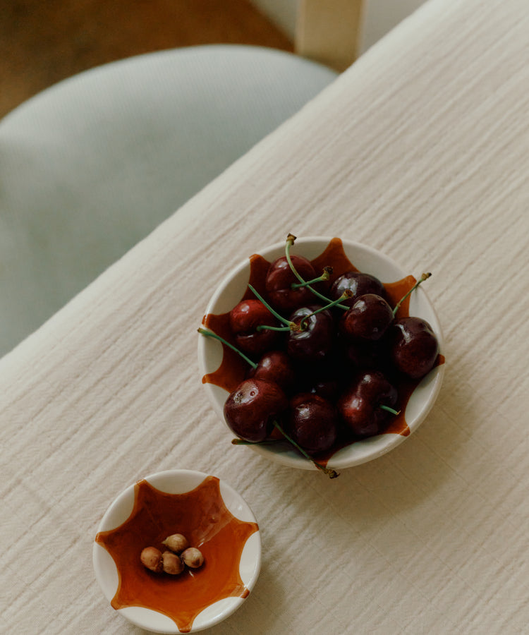 The Stella Nesting Bowls—one filled with cherries and the smaller holding cherry pits—rest on a cream-colored textured tablecloth by a chair, their distinctive ceramics lending artisanal charm to the setting.