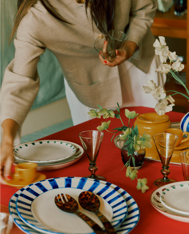 Person setting a table with decorative plates and glasses on a red tablecloth.