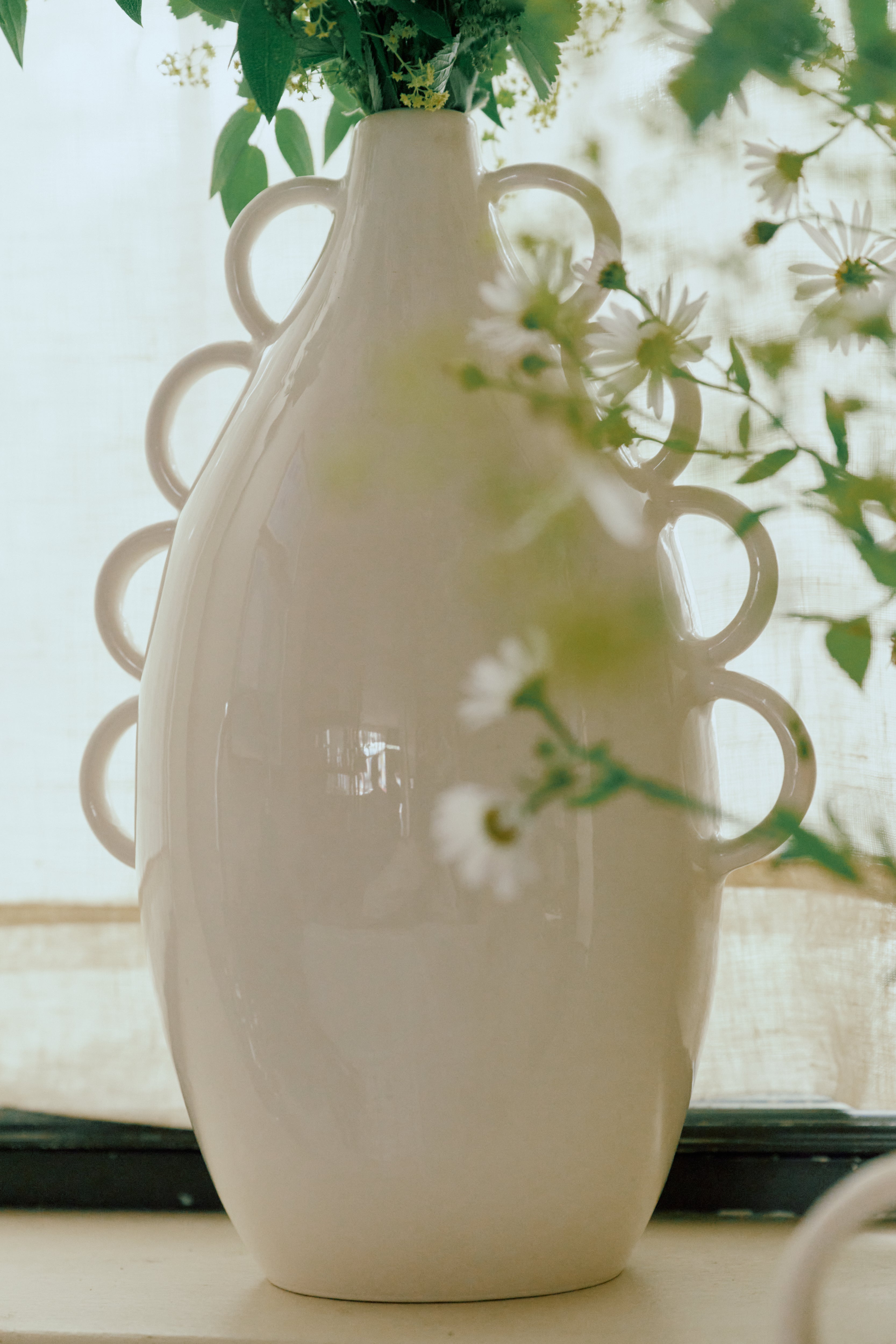 The Brisa Vase—a tall, glossy white sculptural piece featuring decorative looped handles—beautifully displays green foliage and white flowers against a light background, making it the perfect addition to minimal decor or a striking highlight for your modern home accessories collection.