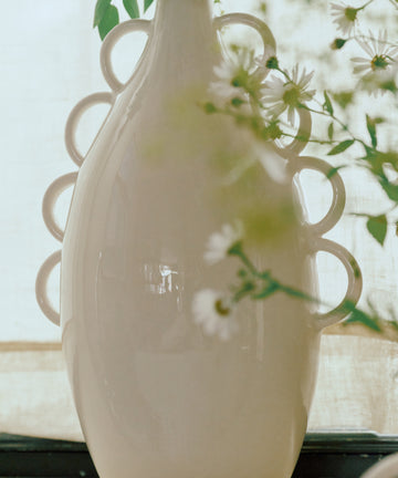 The Brisa Vase—a tall, glossy white sculptural piece featuring decorative looped handles—beautifully displays green foliage and white flowers against a light background, making it the perfect addition to minimal decor or a striking highlight for your modern home accessories collection.