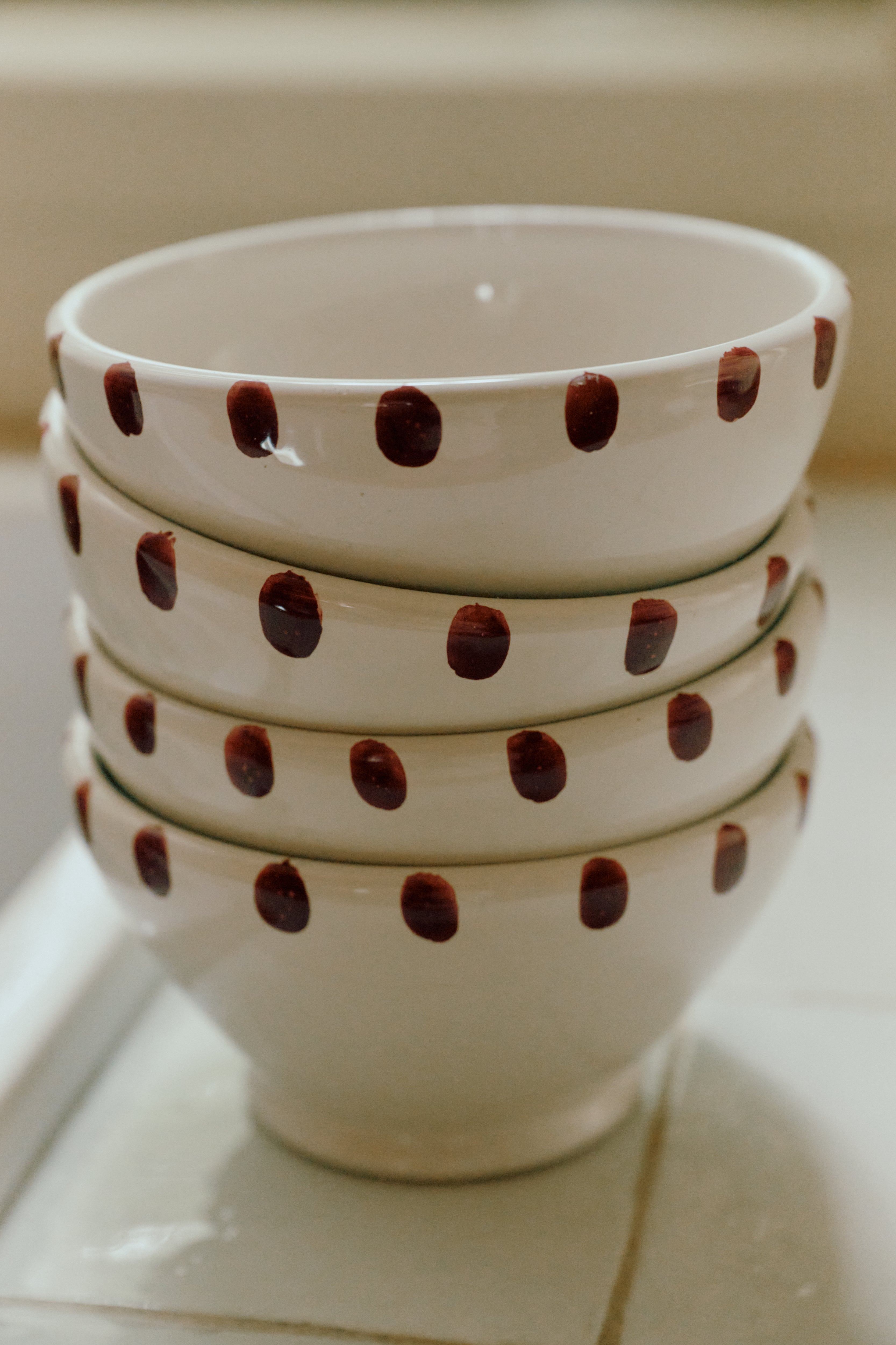 A set of four white ceramic Nina Bowls, each featuring a charming dotted rim and brown polka dots, is stacked neatly on a tiled surface.