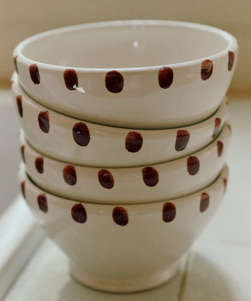 A set of four white ceramic Nina Bowls, each featuring a charming dotted rim and brown polka dots, is stacked neatly on a tiled surface.