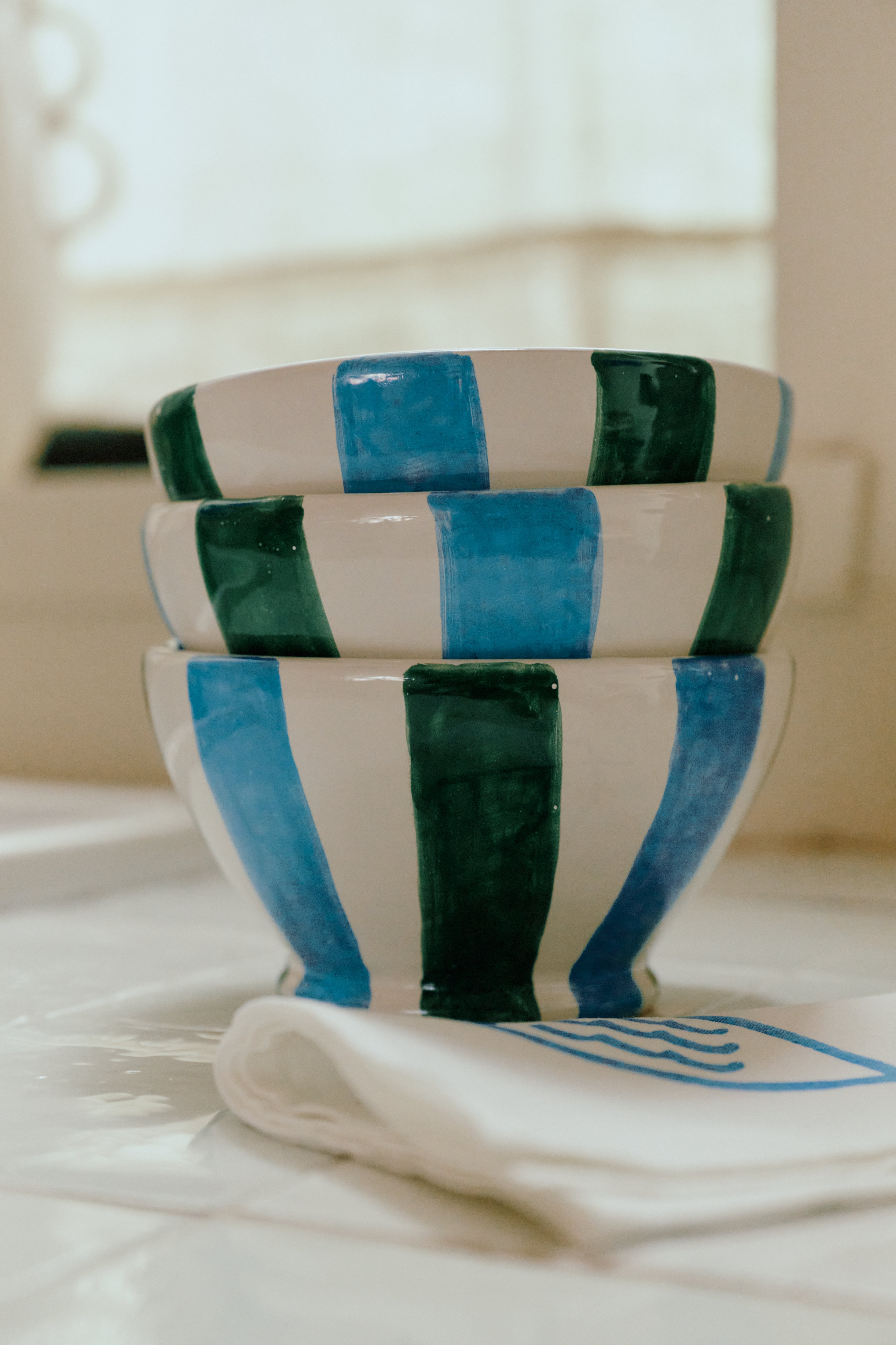 A set of four Mina Bowls, each hand-painted with alternating blue and green vertical stripes, is stacked on a white surface beside a folded napkin, bringing Mediterranean flair to your table.