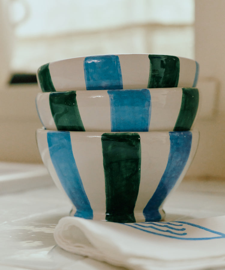 A set of four Mina Bowls, each hand-painted with alternating blue and green vertical stripes, is stacked on a white surface beside a folded napkin, bringing Mediterranean flair to your table.