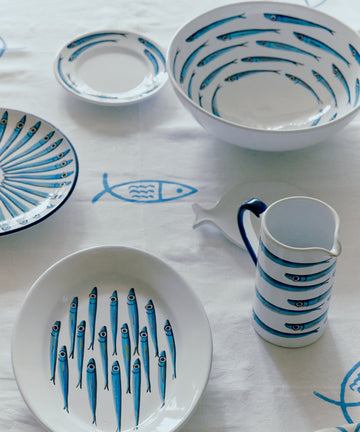 The Tommaso Oval Platter, featuring blue sardine designs, is displayed on a white tablecloth with fish illustrations, capturing the playful coastal charm of the Amalfi Coast.