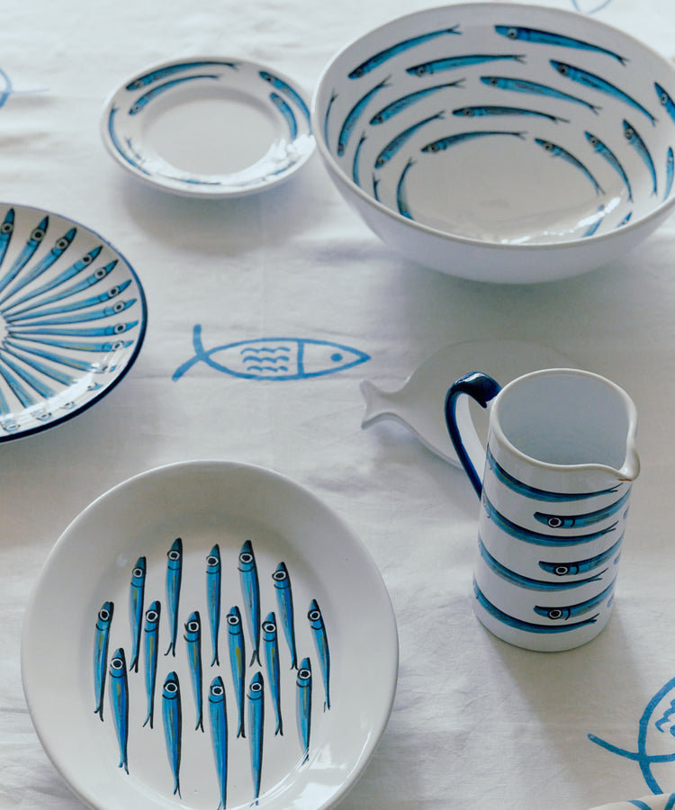 The Tommaso Oval Platter, featuring blue sardine designs, is displayed on a white tablecloth with fish illustrations, capturing the playful coastal charm of the Amalfi Coast.