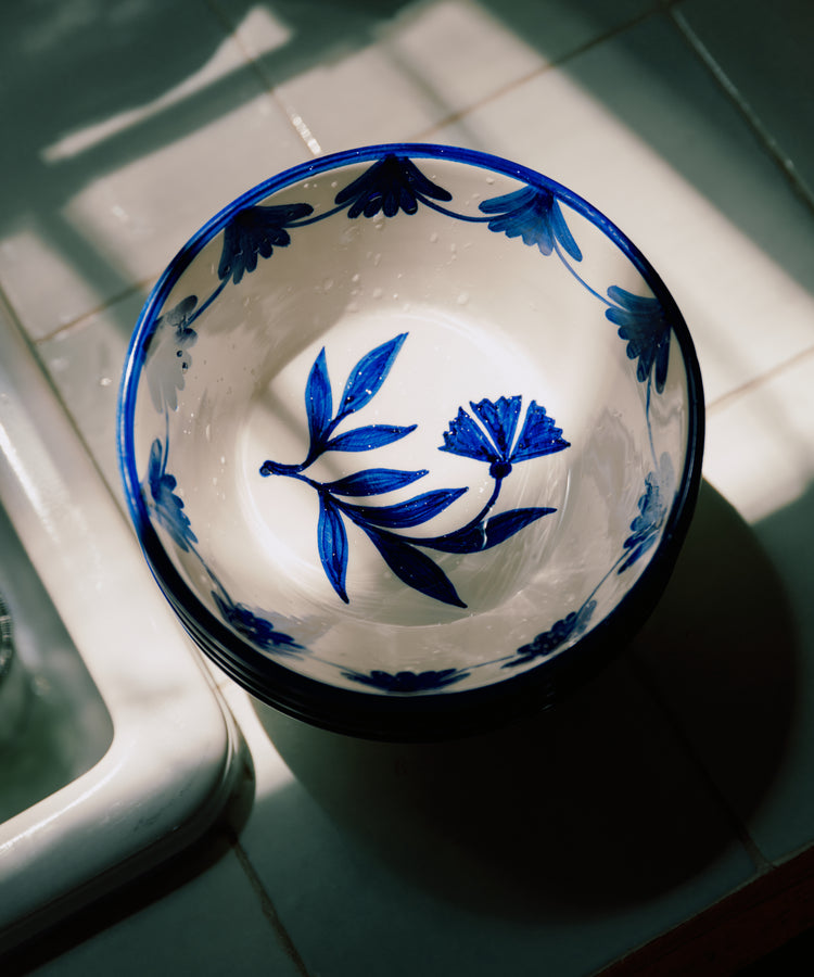 The Catalina Cereal Bowl, Set of 4, features hand-painted ceramic bowls from Portugal with blue floral designs, displayed on a tiled surface and partly lit by sunlight.