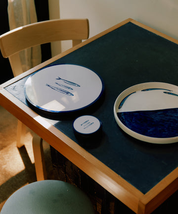 A small wooden table with a wipeable surface is set with Costa Placemats (set of 6) and three round, blue-and-white ceramic plates. A wooden chair and stool nearby create a relaxed summer vibe as sunlight streams in from the left.