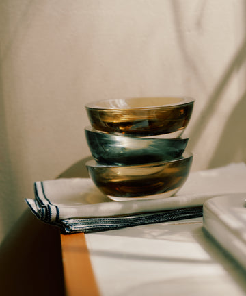 Three Gem Dish bowls in smoke green glass, hand-blown in the South of France, are stacked on folded white napkins with striped edges and placed on a wooden surface in natural light.
