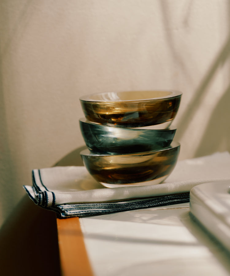 Three Gem Dish bowls in smoke green glass, hand-blown in the South of France, are stacked on folded white napkins with striped edges and placed on a wooden surface in natural light.