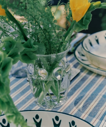 The Carlotta Vase, mouth-blown and filled with yellow flowers and greenery, rests on a blue-and-white striped tablecloth next to patterned ceramic dishes, reflecting the charm of Bohemian artistry.