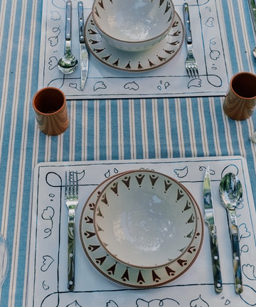 Two place settings on a striped tablecloth with patterned plates, bowls, silverware, and brown cups arranged neatly on wipeable Capriccio Placemats, Set of 6—featuring original decor inspired by PORTA artwork.