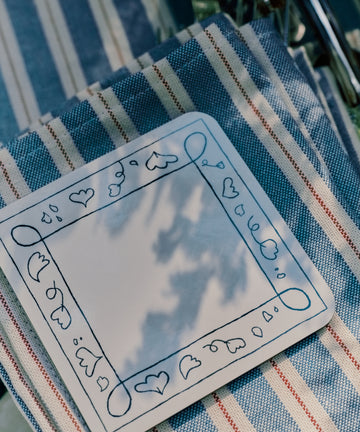 A set of Capriccio Coaster, featuring a playful heart border design on a blank white surface, rests on a striped cloth with blue and red lines; the shadows of leaves cast onto the coasters add to their whimsical appeal.