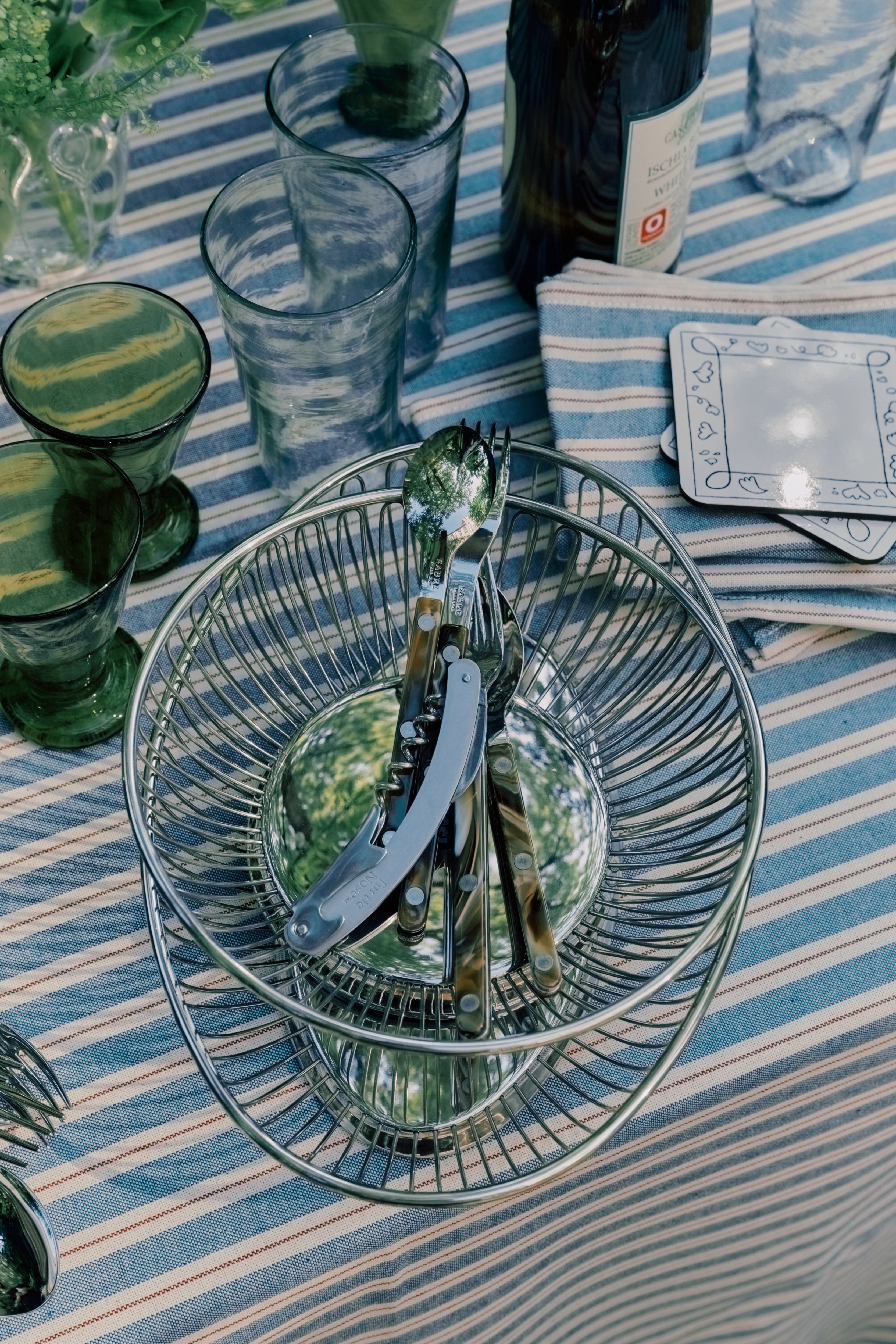 A Round Bread Basket filled with cutlery sits on a striped tablecloth among green glassware, clear glasses, a wine bottle, and a stack of napkins.
