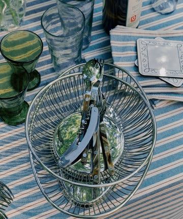 A Round Bread Basket filled with cutlery sits on a striped tablecloth among green glassware, clear glasses, a wine bottle, and a stack of napkins.