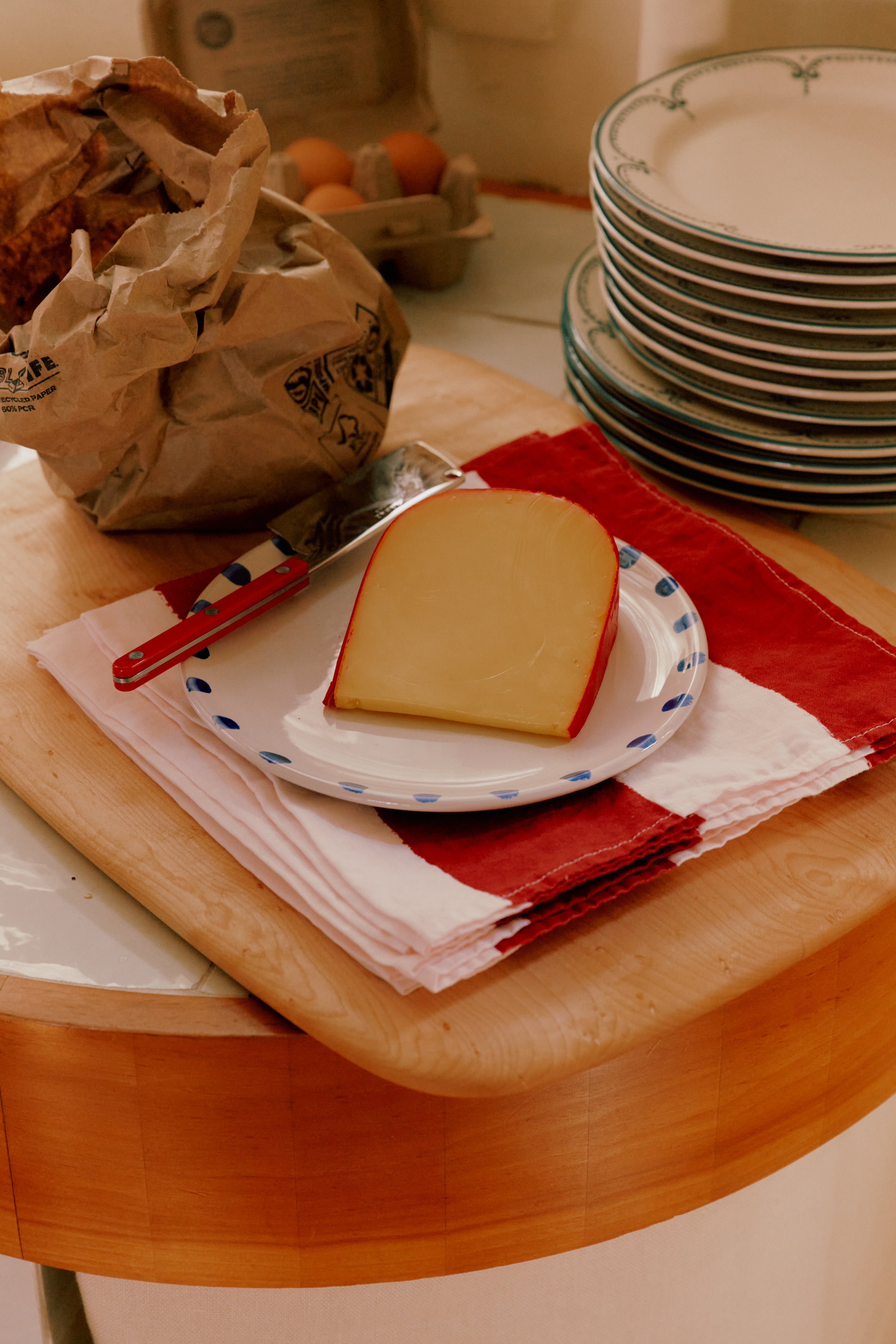A slice of cheese with a knife on painterly table linens, featuring the Striped Napkins, Set of 6, beside stacked plates and a paper bag on a wooden counter.