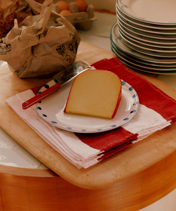 A slice of cheese with a knife on painterly table linens, featuring the Striped Napkins, Set of 6, beside stacked plates and a paper bag on a wooden counter.
