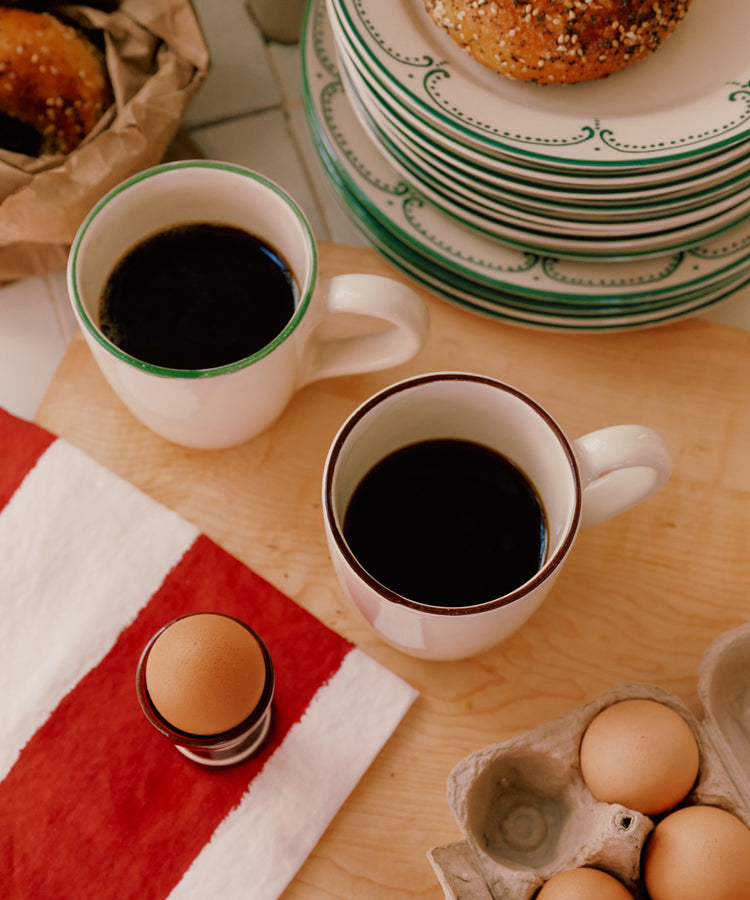 Two Guido Mugs, a soft-boiled egg in a holder, a carton of eggs, a striped napkin, and bagels on plates are set on a wooden table.