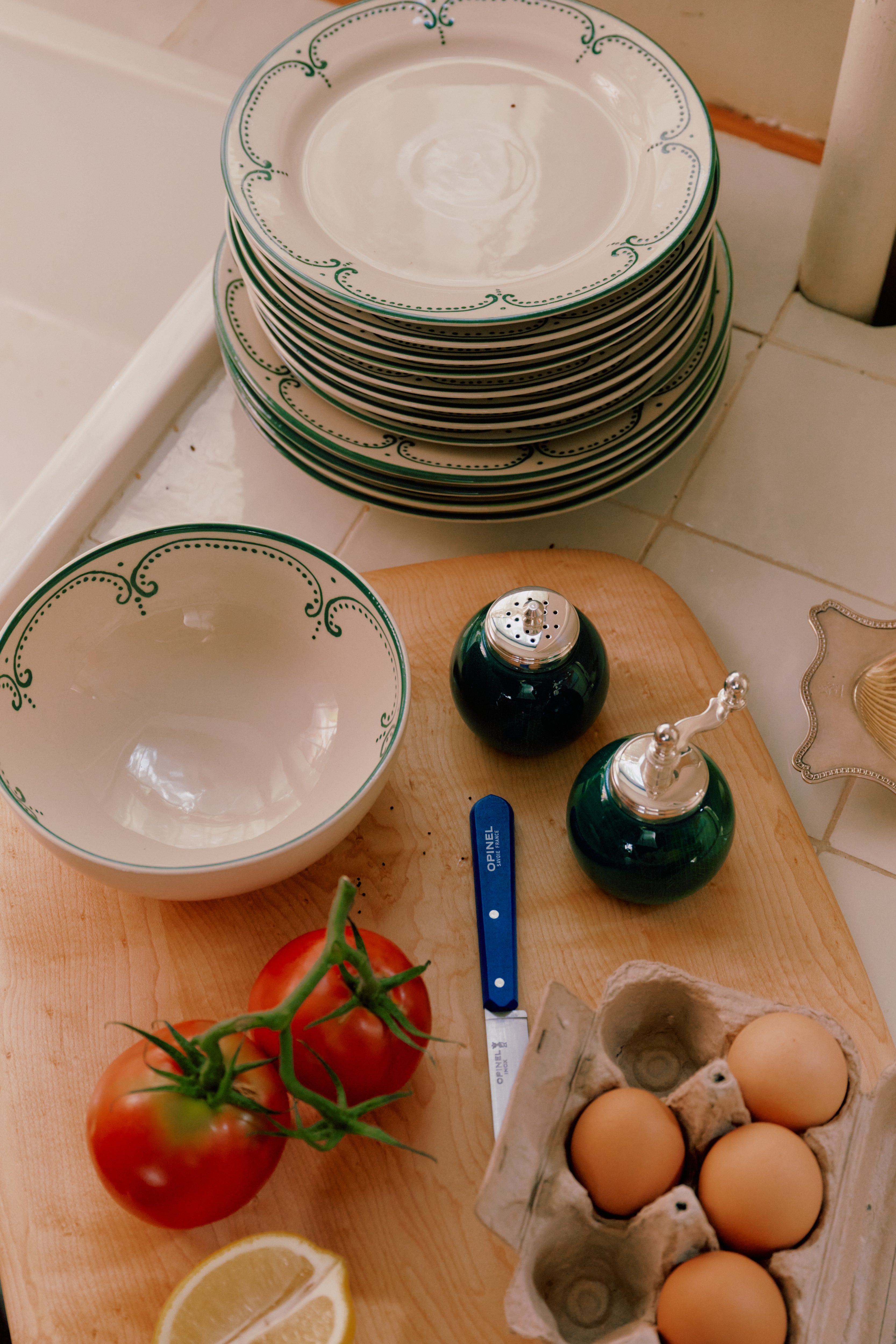 A stack of Arco Dinner Plate, Set of 4, and a bowl rest on a counter beside a cutting board holding tomatoes, a lemon wedge, eggs, a knife, and salt and pepper shakers.