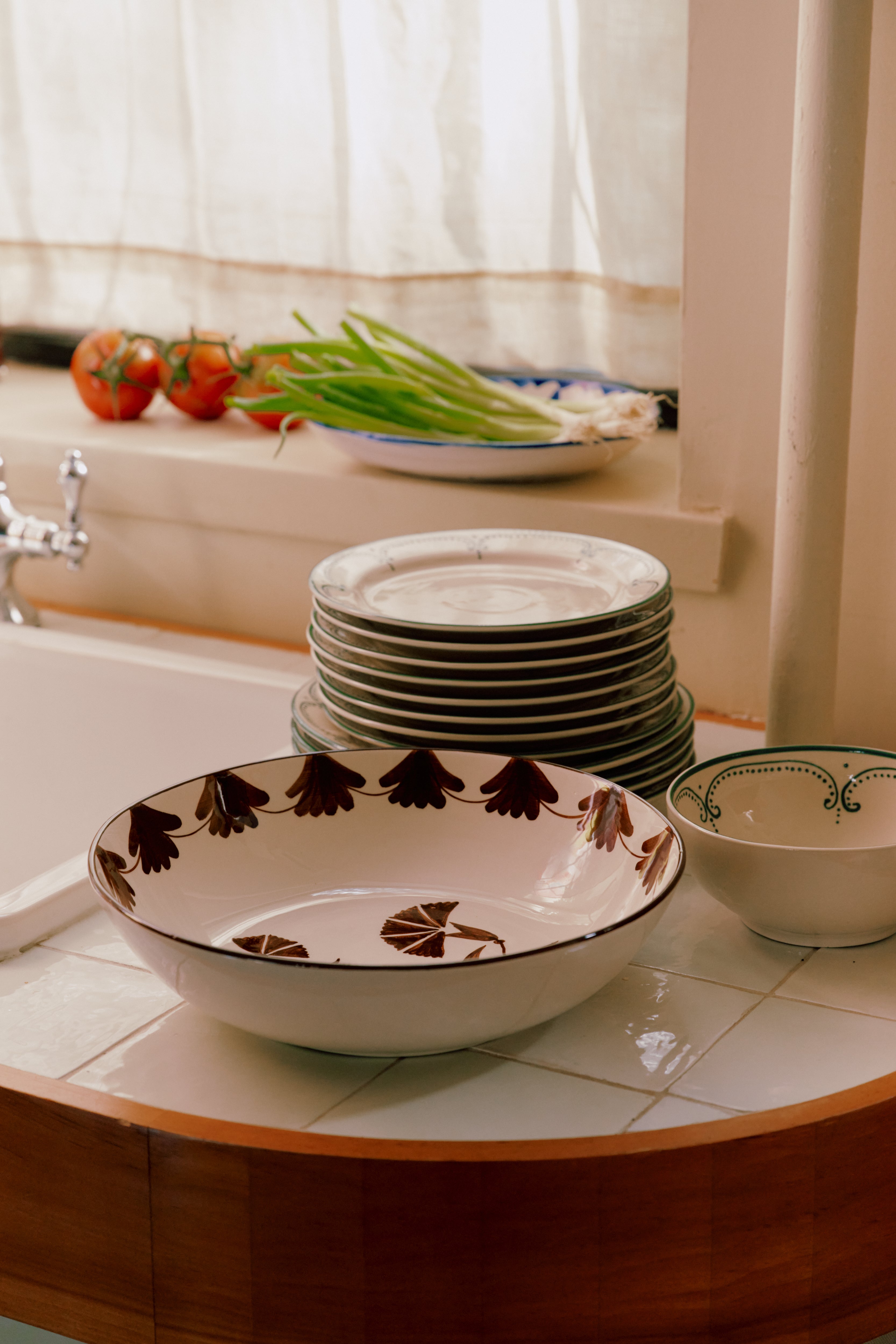 The Catalina Serving Bowl sits on the tiled kitchen counter beside a stack of plates, with tomatoes and green onions arranged nearby and sunlight streaming in through the window.